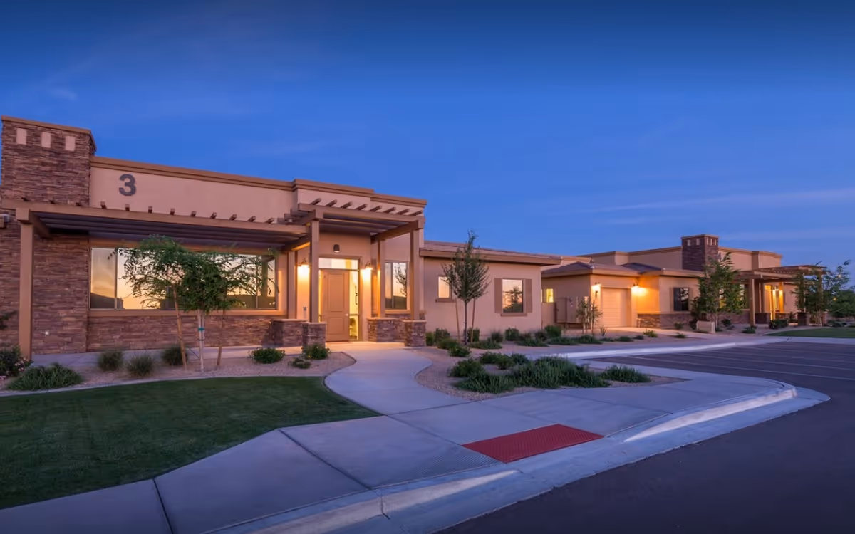 Exterior view of a single-story senior living facility building at dusk with warm lights glowing from inside. The building features stone and stucco walls, large windows, and a covered entrance with the number 3 displayed. There are landscaped bushes, small trees, and a sidewalk leading to the entrance, with a parking lot visible on the right.