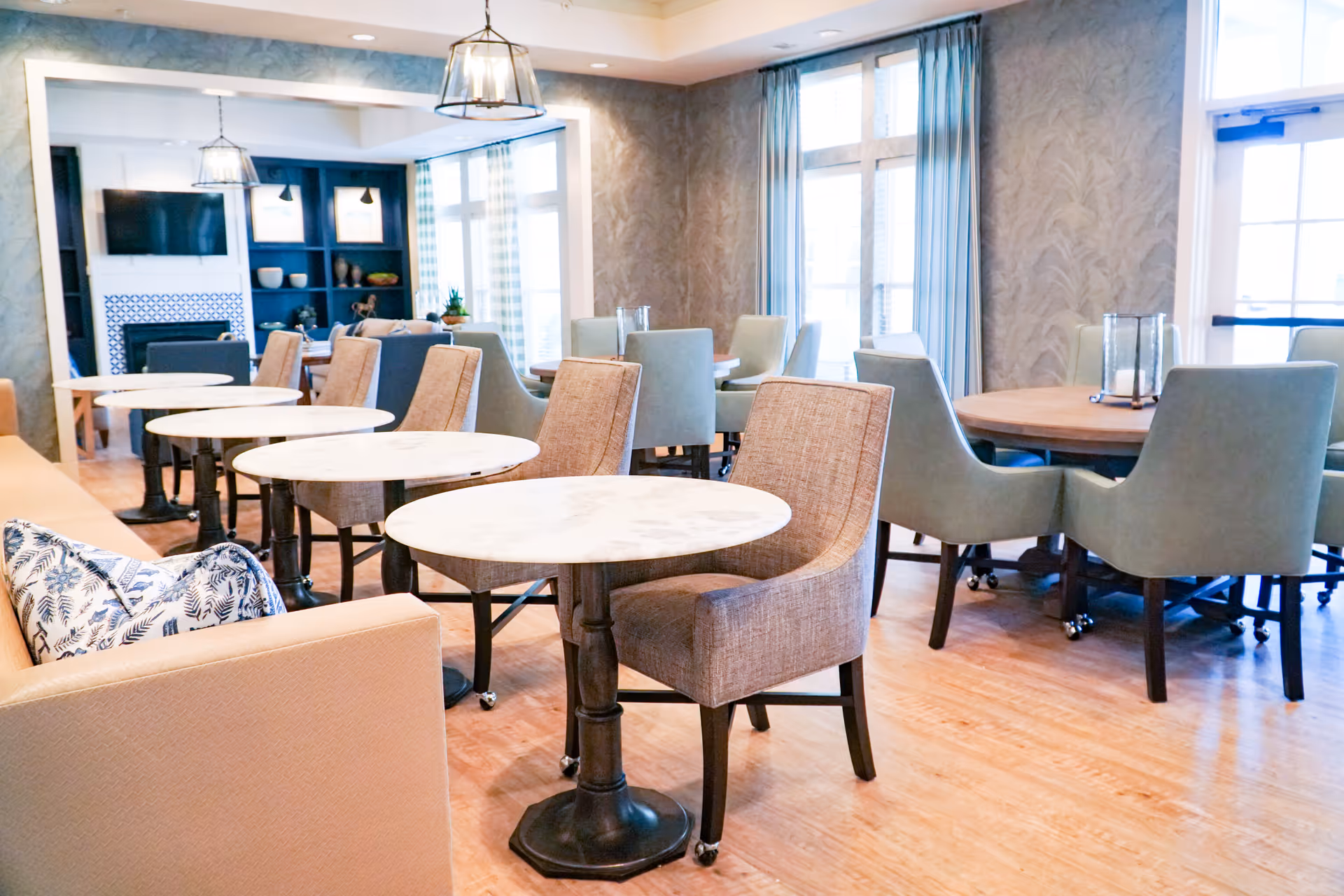 A bright and spacious dining area with multiple round tables and upholstered chairs. The room features large windows with blue curtains, wooden flooring, and a cozy fireplace with a TV mounted above it. Decorative shelves and pendant lights add to the inviting atmosphere.