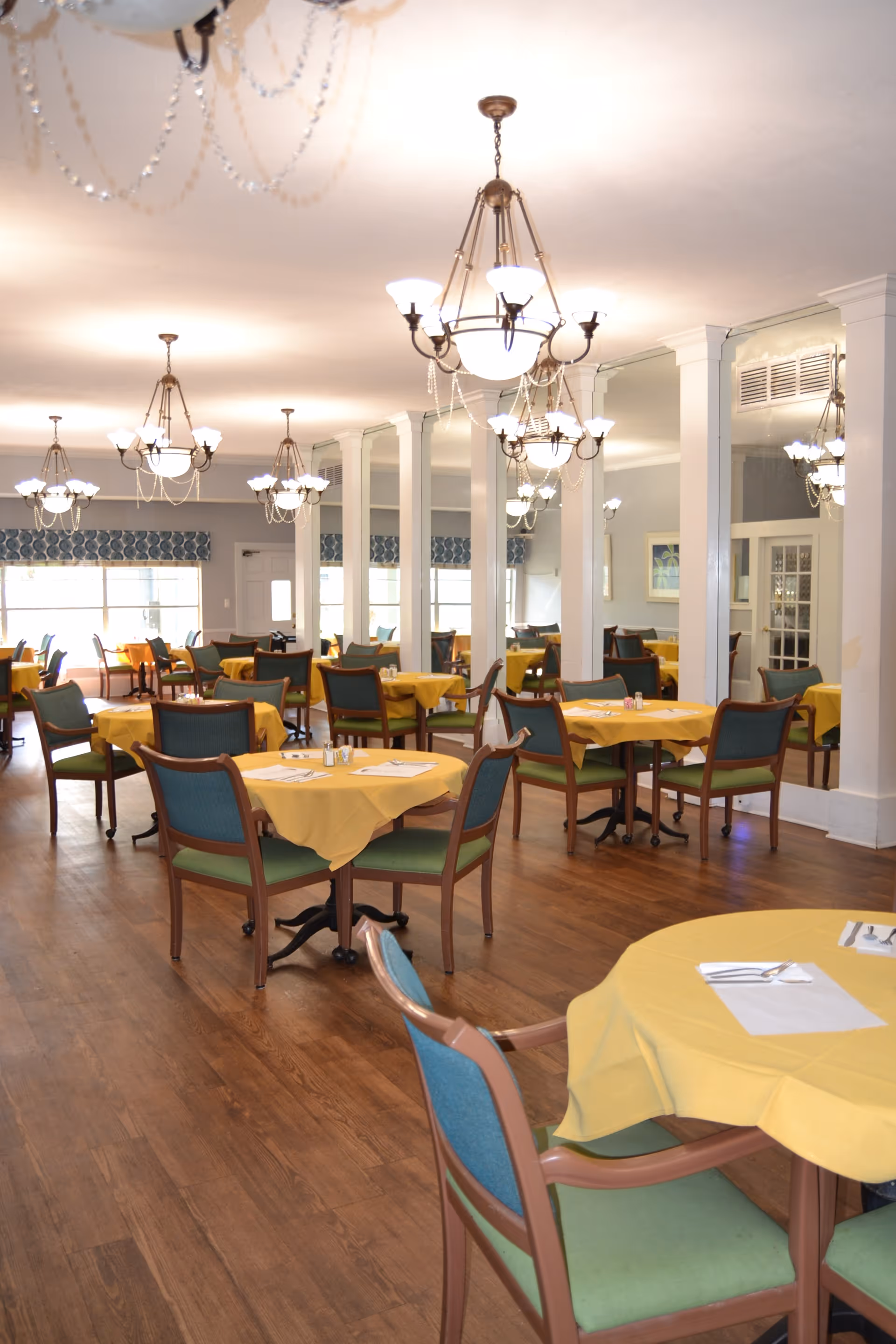 Bright communal dining room with tables covered in yellow tablecloths and green-cushioned chairs under multiple chandeliers.