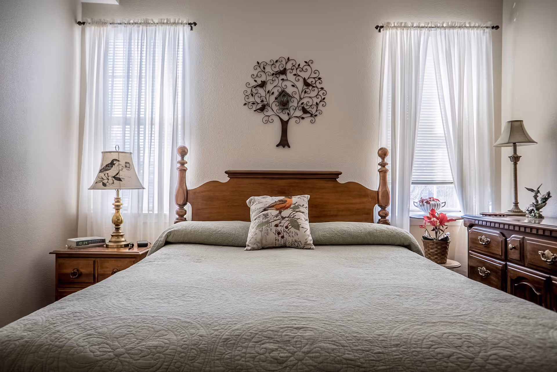 A cozy bedroom with a wooden bed frame and a light green quilted bedspread. There is a decorative pillow with a bird design in the center of the bed. On either side of the bed are wooden nightstands with lamps; the left lamp has a bird design on the shade. Two windows with white sheer curtains flank the bed. Above the bed is a decorative metal wall art piece shaped like a tree with birds.