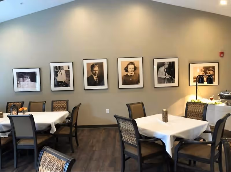 Dining room with tables covered in white tablecloths and chairs arranged around them. The walls are decorated with framed black and white photographs of people. There is a buffet or serving area with food and heat lamps on the right side.