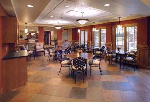 Sunlit dining hall with round tables, chairs, and large windows in a warm-modern facility.