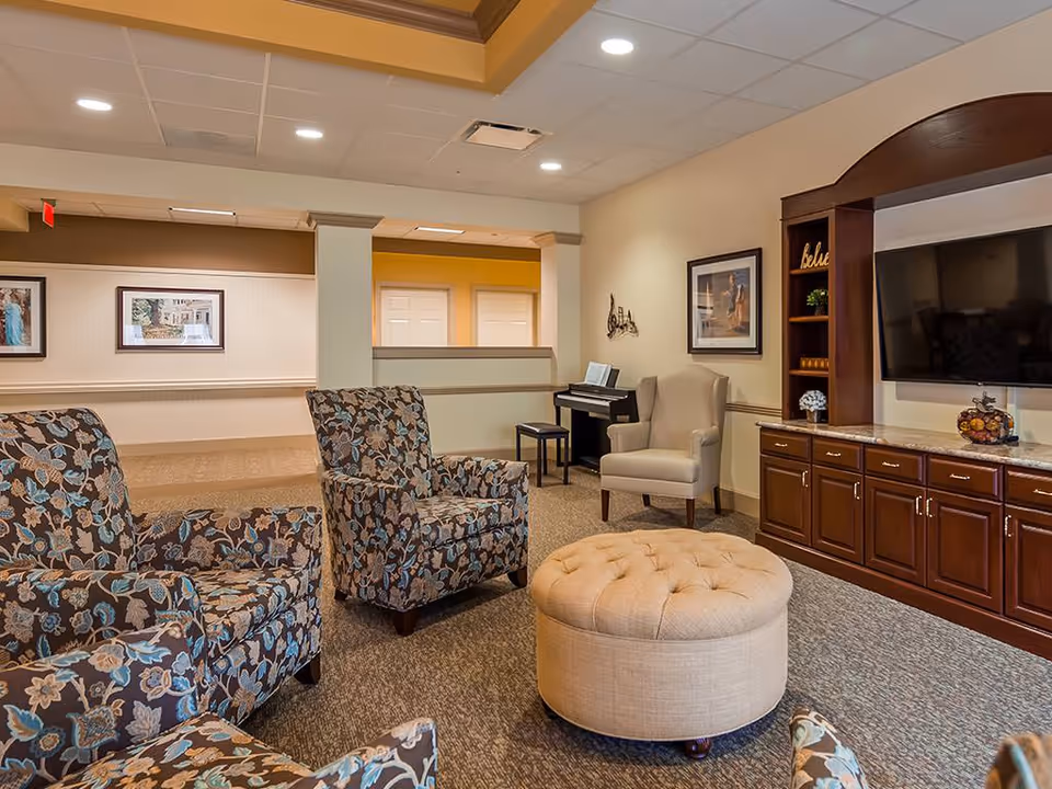 A cozy living room area in a senior living facility featuring three floral-patterned armchairs, a beige tufted ottoman, a beige wingback chair, a digital piano with a stool, and a large flat-screen TV mounted above a wooden cabinet with shelves and drawers. The walls are decorated with framed pictures and a decorative wall piece, and the room has a carpeted floor and recessed lighting in the ceiling.