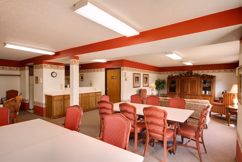 Communal dining and sitting area with red upholstered chairs, tables, and wooden cabinetry in a senior living facility.