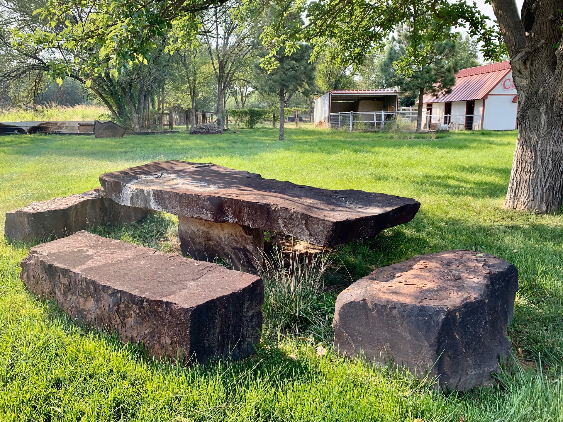 Outdoor area with a rustic stone table and three stone benches on green grass under trees, with a barn and fenced area in the background.