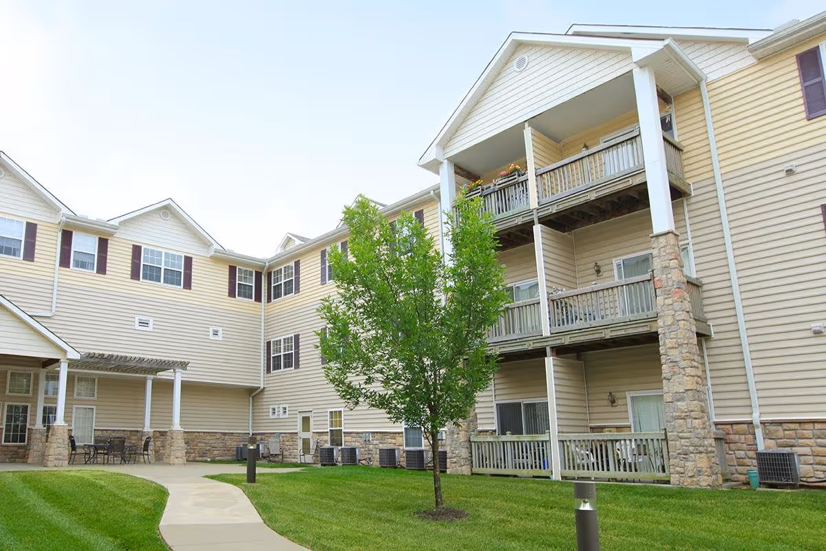 Exterior view of a multi-story senior living facility building with beige siding and stone accents. The building has balconies with railings and a small tree on a well-maintained lawn with a curved concrete pathway. There is outdoor seating under a covered area on the left side.