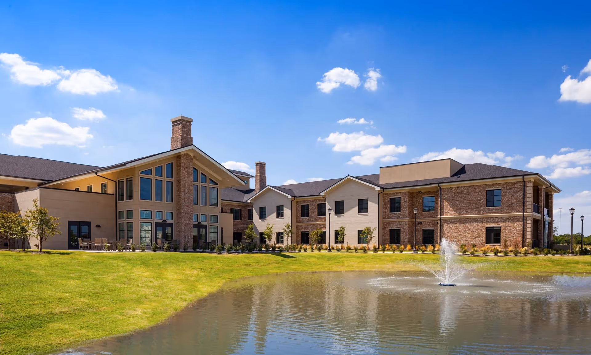 Exterior view of HarborChase of Plano senior living facility showing a large two-story building with brick and beige walls, multiple windows, and chimneys. The building is situated next to a pond with a water fountain, surrounded by a well-maintained green lawn under a blue sky with scattered clouds.