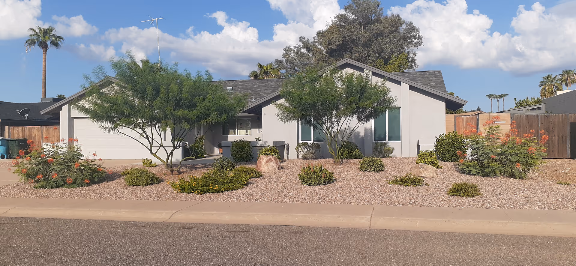 Single-story house front with attached garage, desert rock landscaping and small trees under a partly cloudy sky.