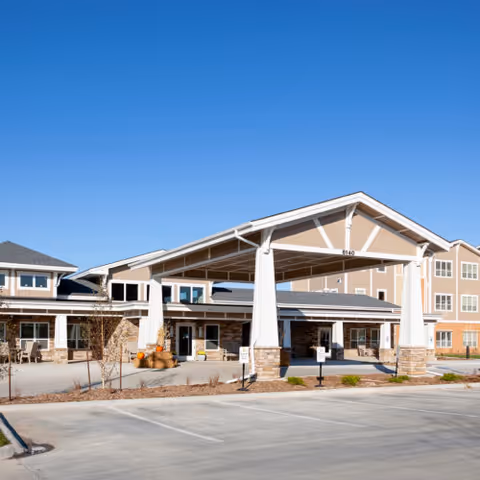 Exterior view of Cedarhurst Senior Living of La Vista showing the main entrance with a large covered drop-off area supported by white columns. The building has multiple windows and a combination of stone and beige siding under a clear blue sky.