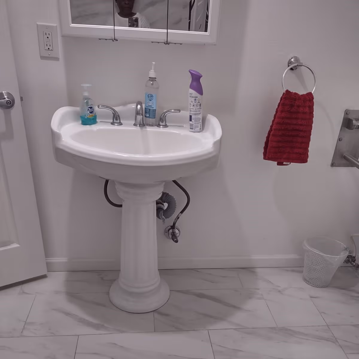 A white pedestal sink in a bathroom with a mirror above it. On the sink are a bottle of hand soap, a bottle of hand sanitizer, and a spray bottle. To the right of the sink is a red hand towel hanging on a silver towel ring and a small white trash bin on the floor. The floor is tiled with white and gray marble pattern.