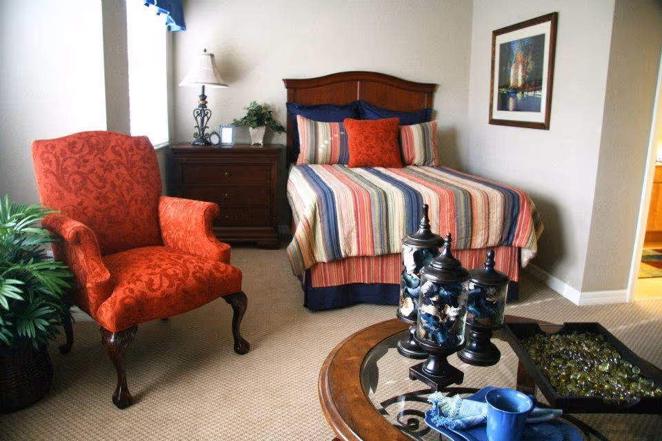 A cozy bedroom featuring a bed with a striped multicolored bedspread and multiple pillows, a wooden nightstand with a lamp and plant, a red upholstered armchair, a glass-top coffee table with decorative jars, and a framed painting on the wall.