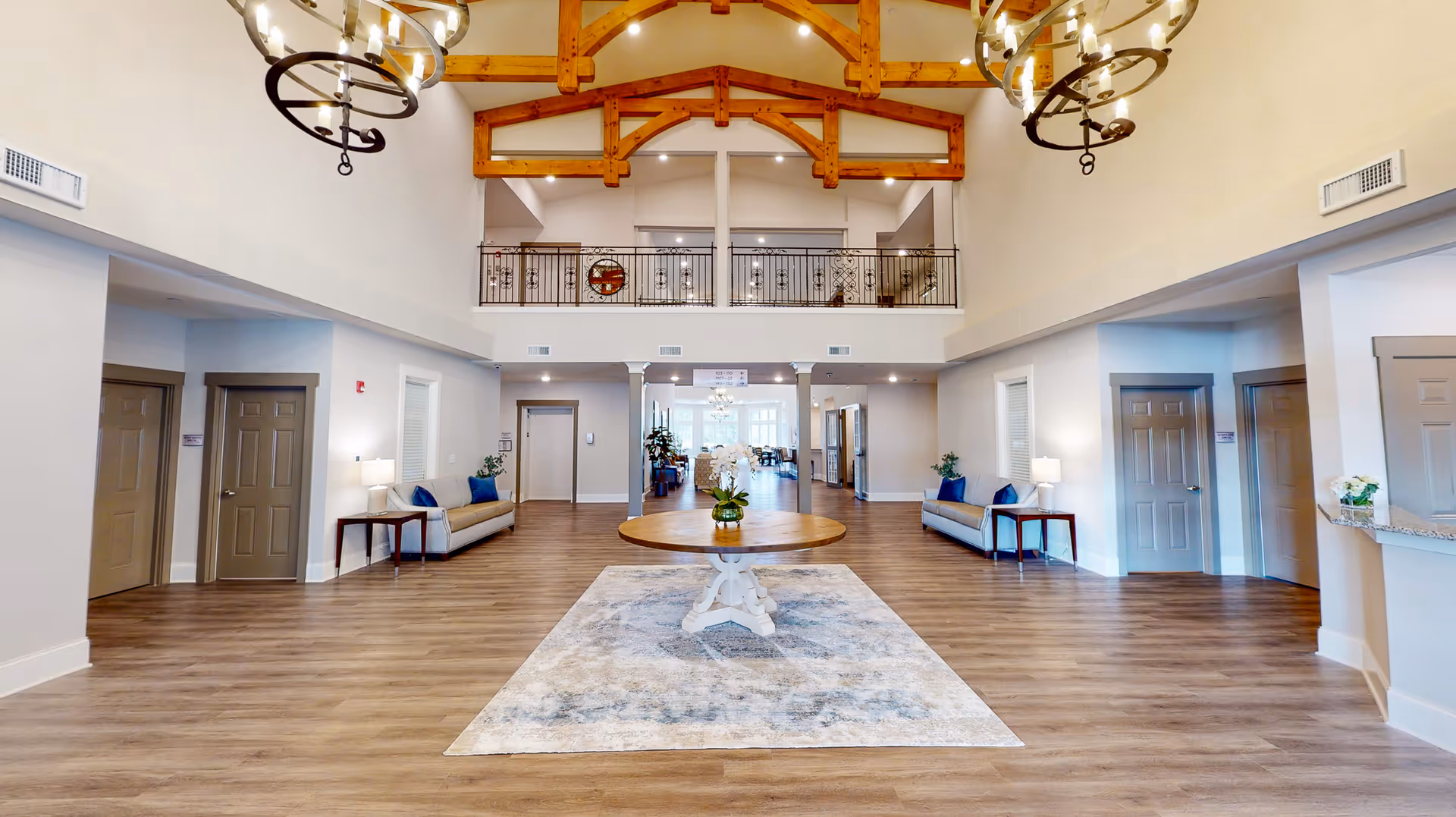 Spacious two-story senior living lobby with exposed wooden beams, a central round table on a rug, seating along the walls, and hanging chandeliers.