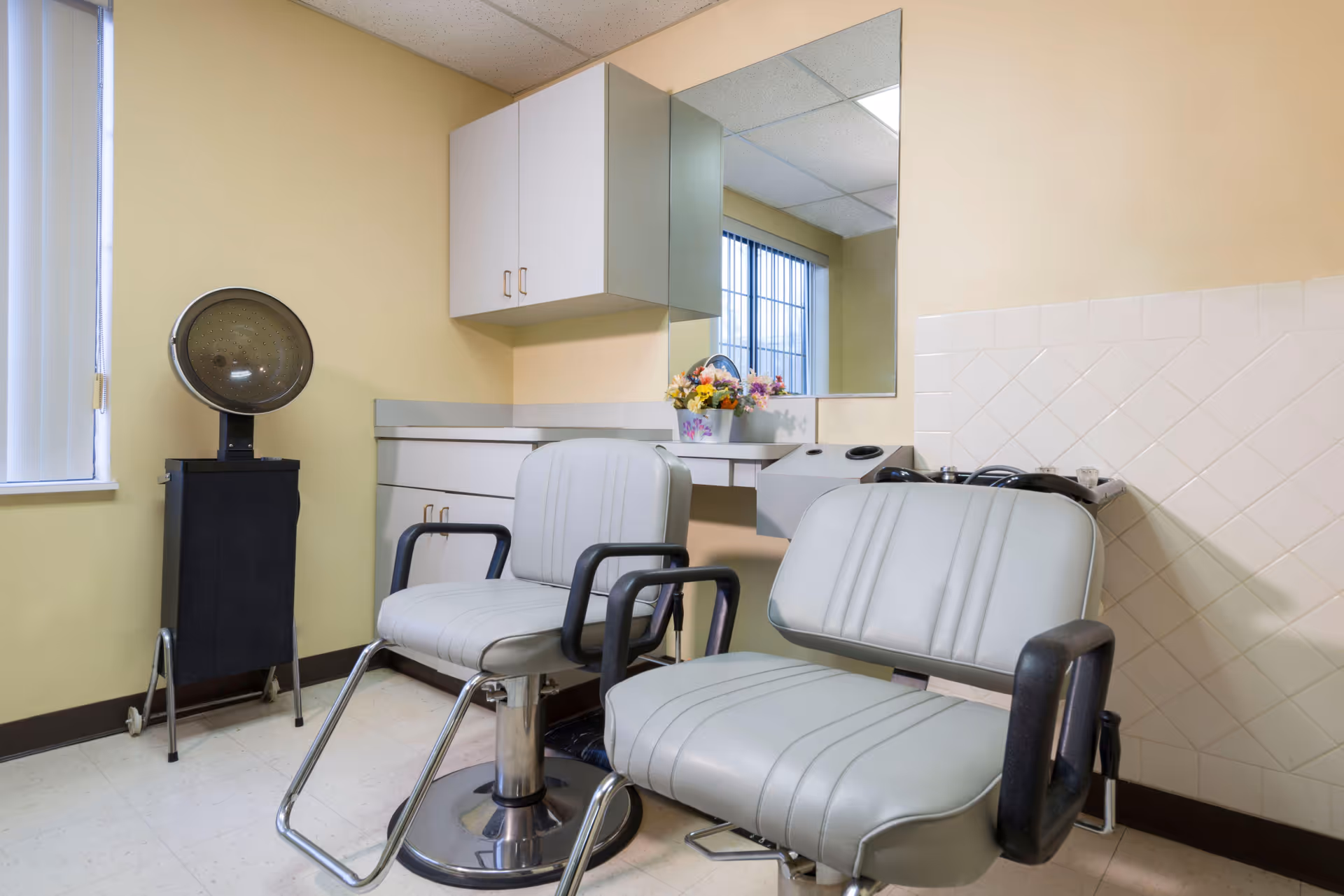 A small salon room with two light gray salon chairs with black armrests, a vintage hair dryer on a stand, a countertop with cabinets above and below, a large mirror, and a window with vertical blinds. There is a small floral arrangement on the countertop.