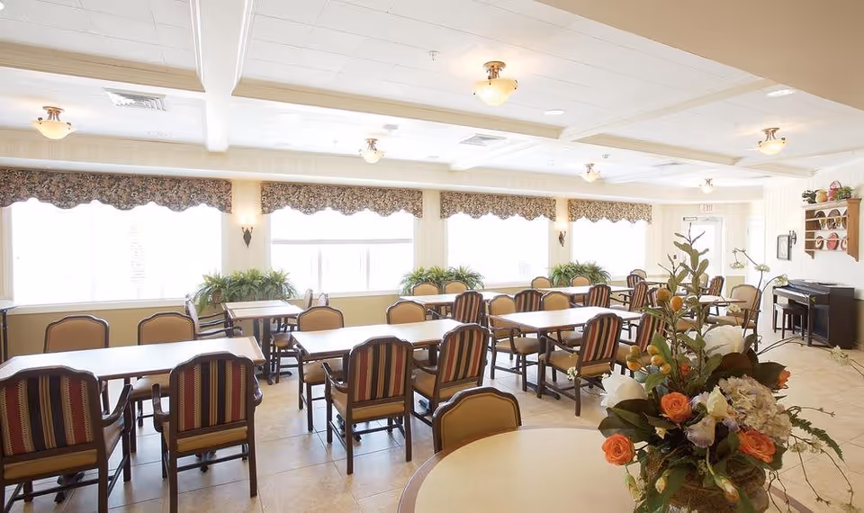 A bright dining room with multiple tables and chairs arranged neatly. The room has large windows with floral valances allowing natural light to fill the space. There are plants on the windowsills and a floral arrangement on a round table in the foreground. A piano and decorative shelves are visible in the background.