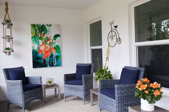 A cozy outdoor seating area with three wicker chairs featuring dark blue cushions arranged around two small tables. The space is decorated with a hanging planter with succulents, a colorful flamingo painting on the wall, a metal wall art bicycle, and potted flowers on the tables. Two windows with blinds are visible on the white wall behind the chairs.