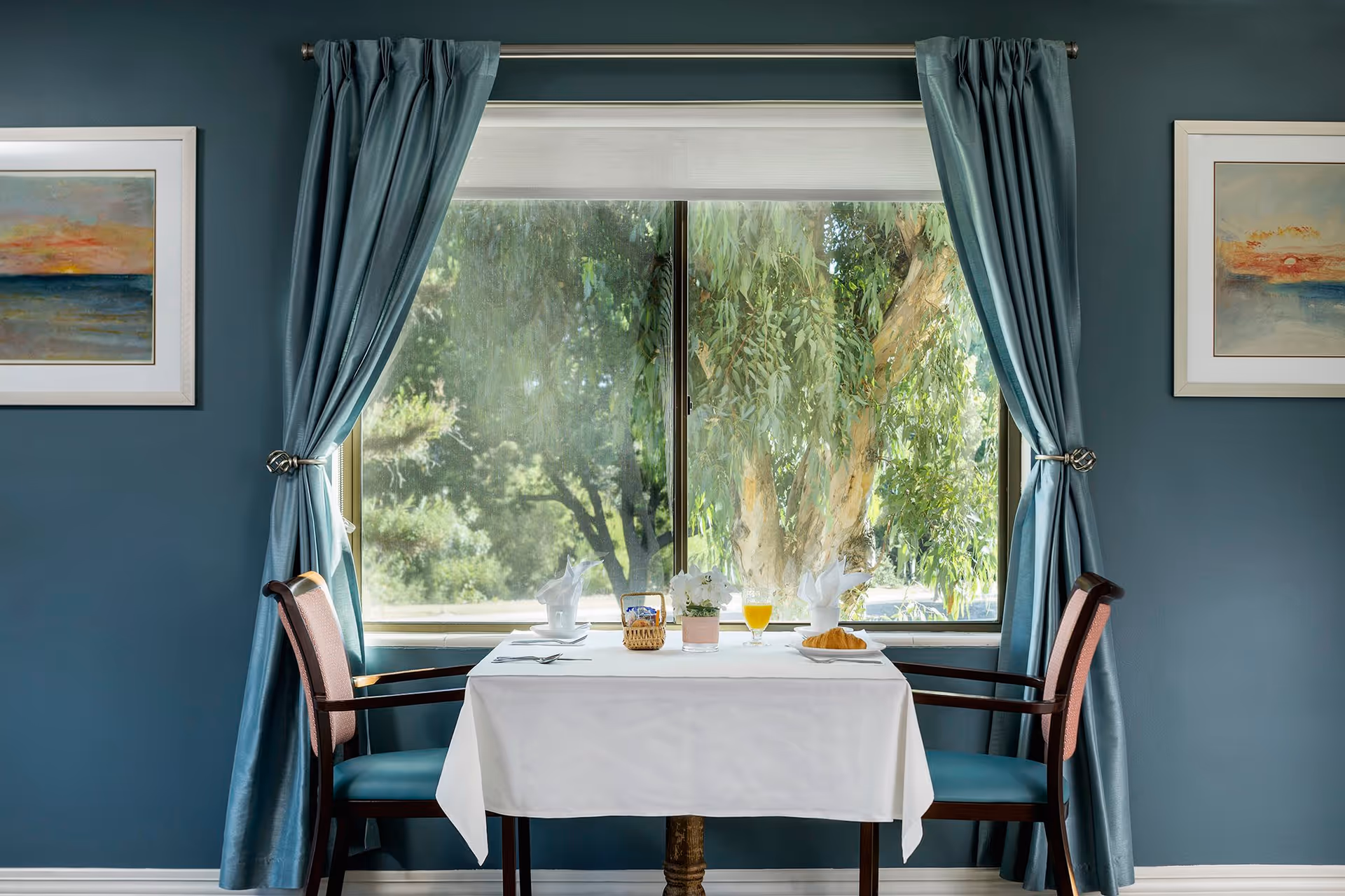 A small dining table set for two with white tablecloth, napkins, a glass of orange juice, a croissant, and a small plant, positioned in front of a large window with blue curtains tied back, showing green trees outside. Two wooden chairs with blue cushions are placed on either side of the table. The walls are painted blue and decorated with two framed paintings of sunsets.