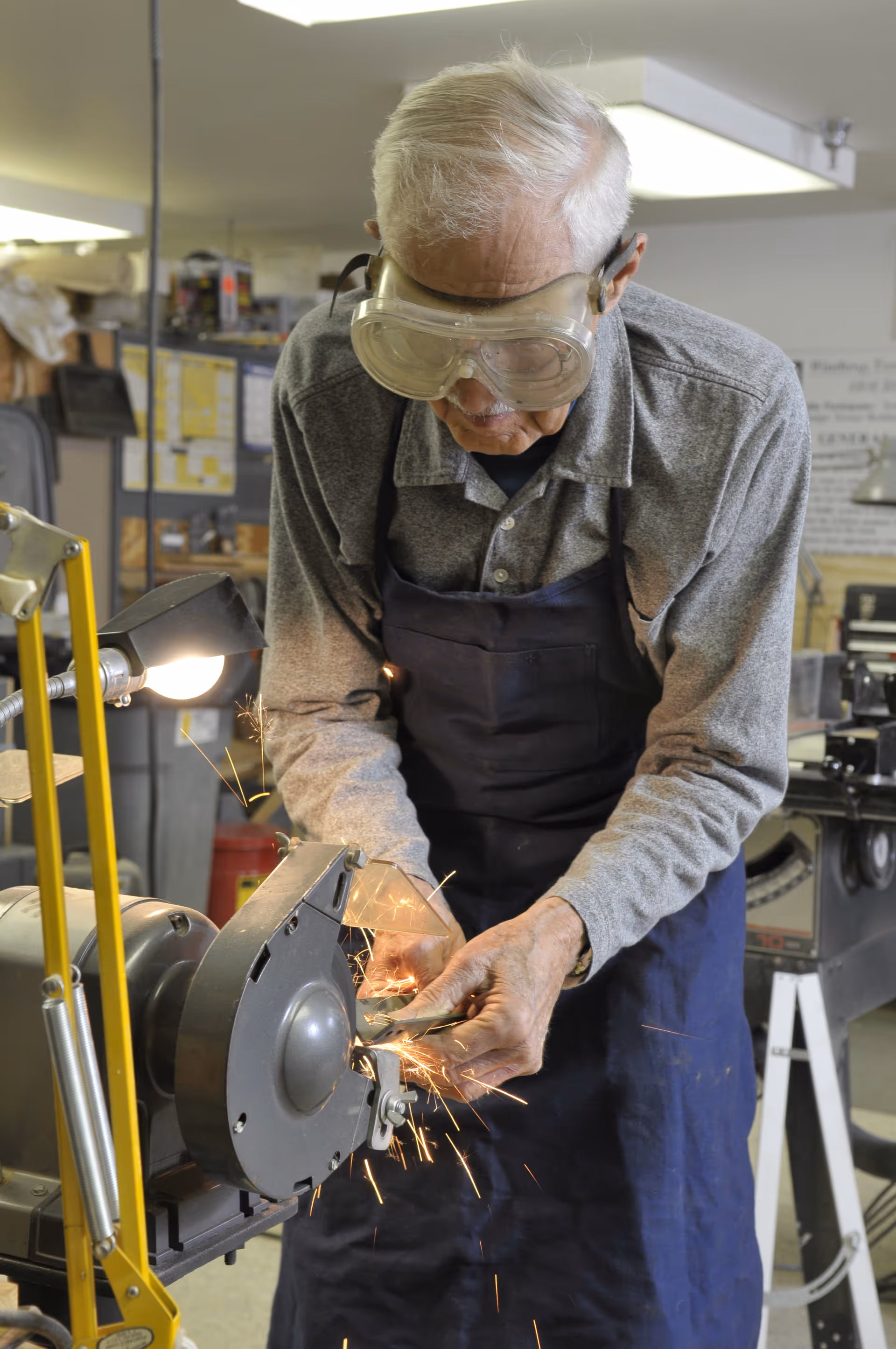 An elderly man wearing safety goggles and a blue apron is using a grinding machine, producing sparks in a workshop setting.