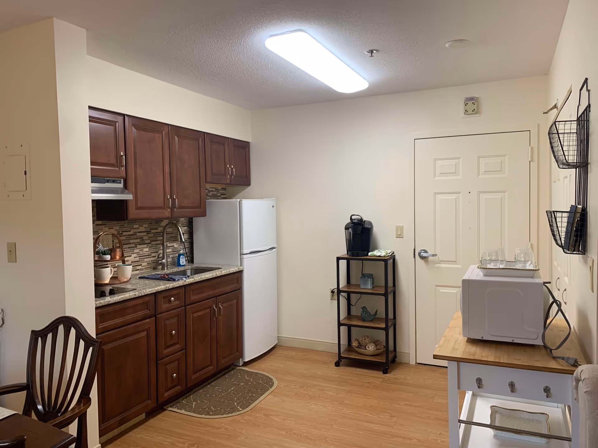 A small kitchen area in a senior living facility with wooden cabinets, a granite countertop, a white refrigerator, a sink, and a stovetop. There is a small shelving unit with a coffee maker and some decorative items. A microwave sits on a wooden table with hooks underneath, and there are wall-mounted wire baskets on the right wall. The floor is wood, and the walls are painted light beige.