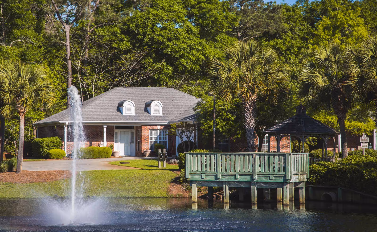 A serene outdoor scene at The Lakes at Litchfield featuring a small pond with a water fountain, a wooden deck extending over the water, palm trees, and a brick building with a gray roof in the background surrounded by lush green trees and shrubs.