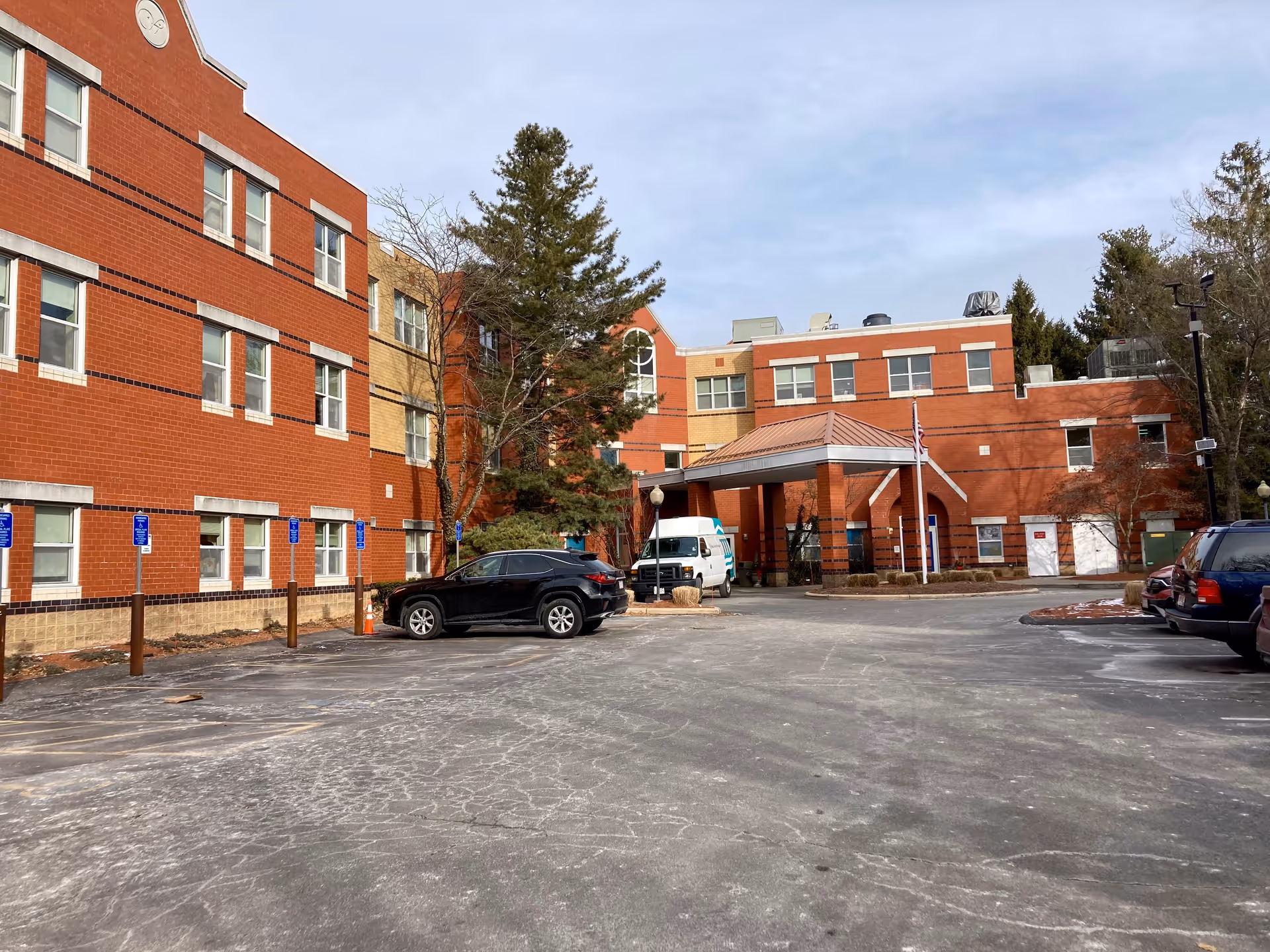Exterior view of a multi-story brick building with a covered entrance and a parking lot in front. Several cars are parked, and there are trees around the building under a partly cloudy sky.