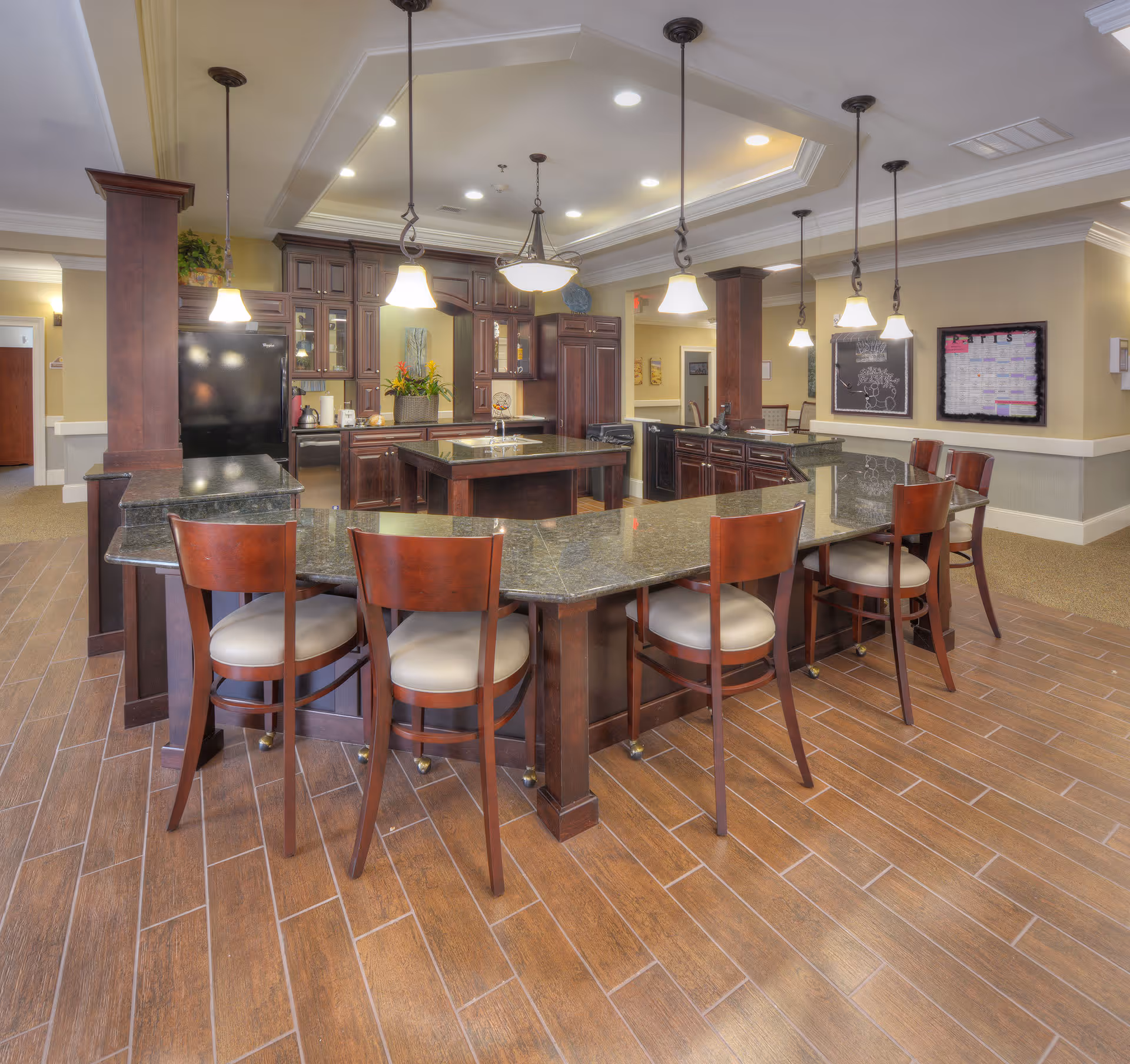 A spacious kitchen area with dark wood cabinetry and a large granite countertop island surrounded by wooden chairs with cushioned seats. The kitchen features pendant lighting hanging from a recessed ceiling with multiple light fixtures. The floor is covered with wood-patterned tiles, and there are decorative plants and framed artwork on the walls.