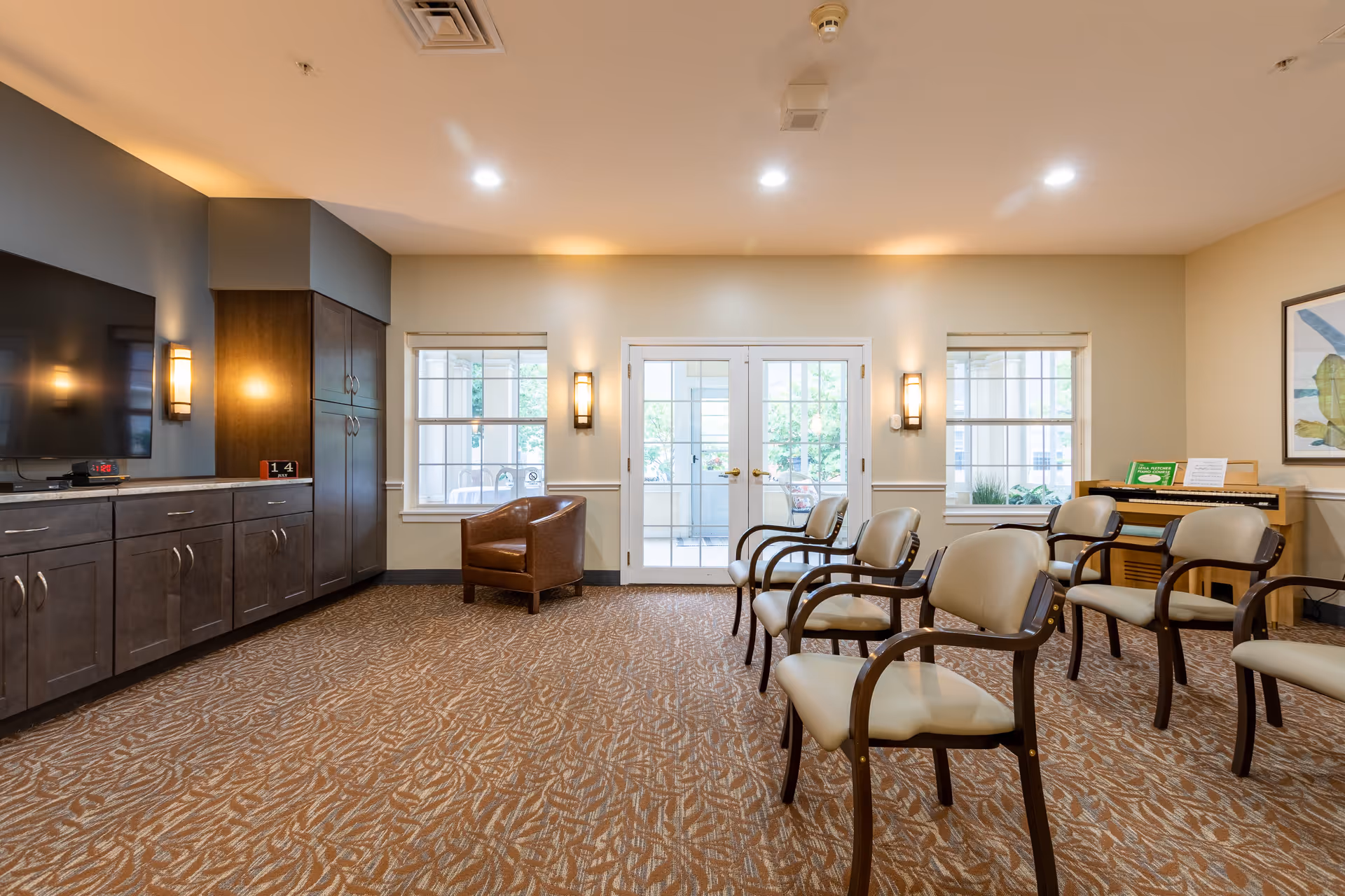 A well-lit common area with several beige cushioned chairs arranged in rows on a patterned carpet. There is a brown leather armchair near two windows and a set of double glass doors leading outside. On the left wall, there is a large flat-screen TV mounted above dark wooden cabinets. On the right side, there is an upright piano with sheet music and a framed artwork on the wall.