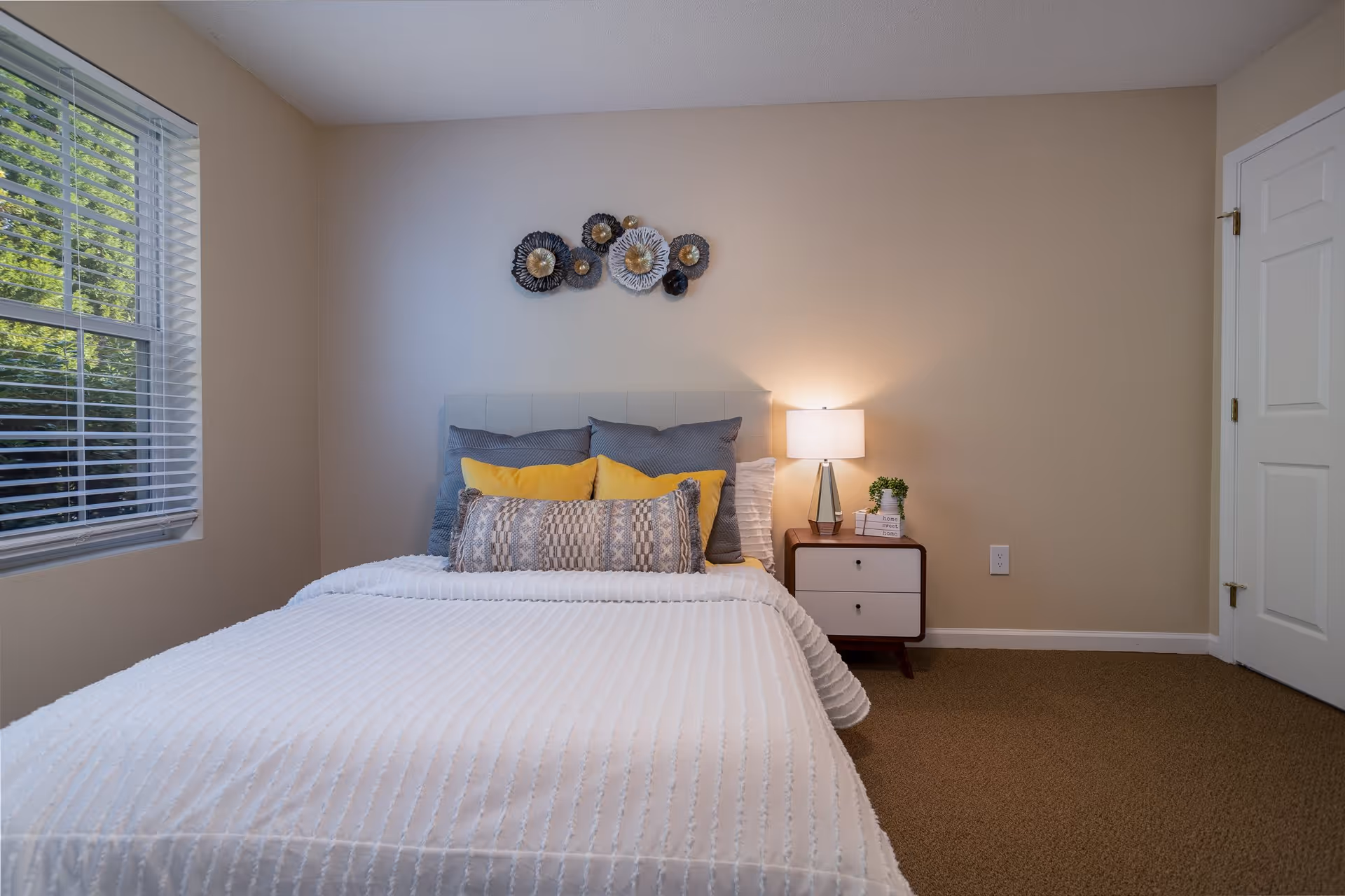 A neatly made bed with white textured bedding and multiple pillows in gray, yellow, and patterned designs. There is a small nightstand with a lamp, a small plant, and books next to the bed. The room has beige walls, a window with white blinds on the left, and a closed white door on the right. Decorative wall art featuring circular metal designs is hung above the bed.