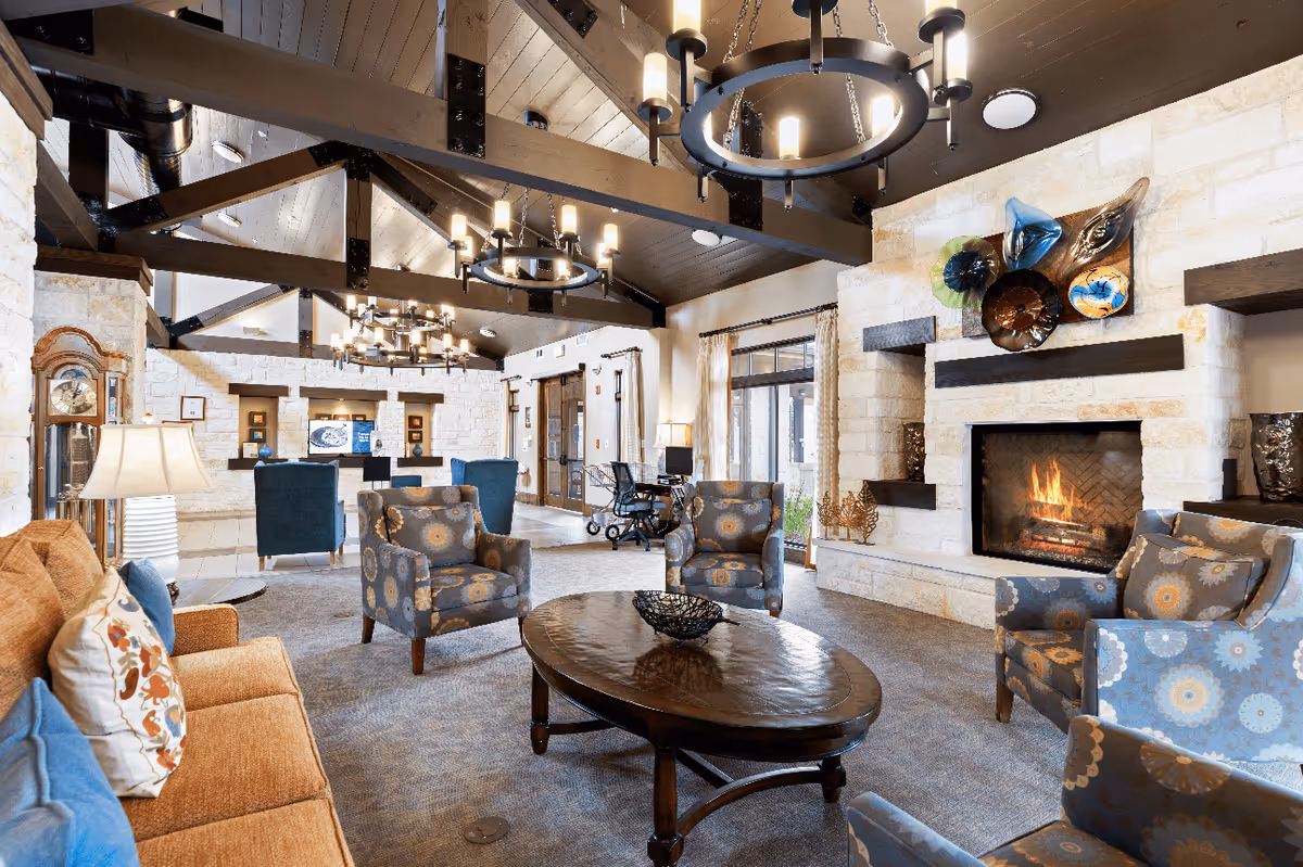 A spacious and well-lit living room area with a high vaulted ceiling featuring exposed wooden beams and multiple circular chandeliers. The room includes a stone fireplace with decorative art above it, several patterned armchairs, a brown sofa with colorful pillows, a wooden coffee table, and a grandfather clock. Large windows with curtains allow natural light to enter the room.
