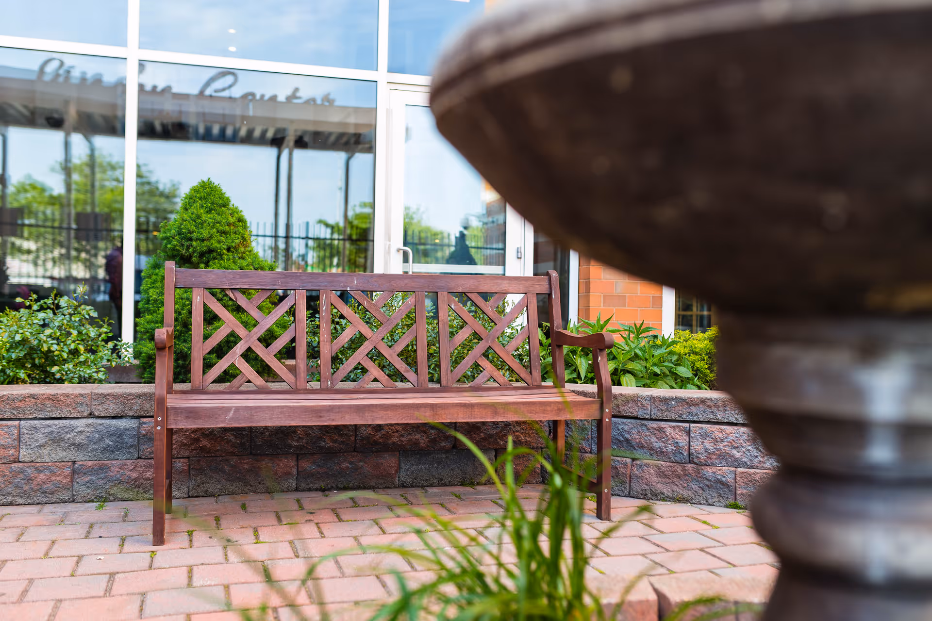 A wooden bench sits on a brick patio in front of a nursing center entrance with plants and a blurred fountain in the foreground.
