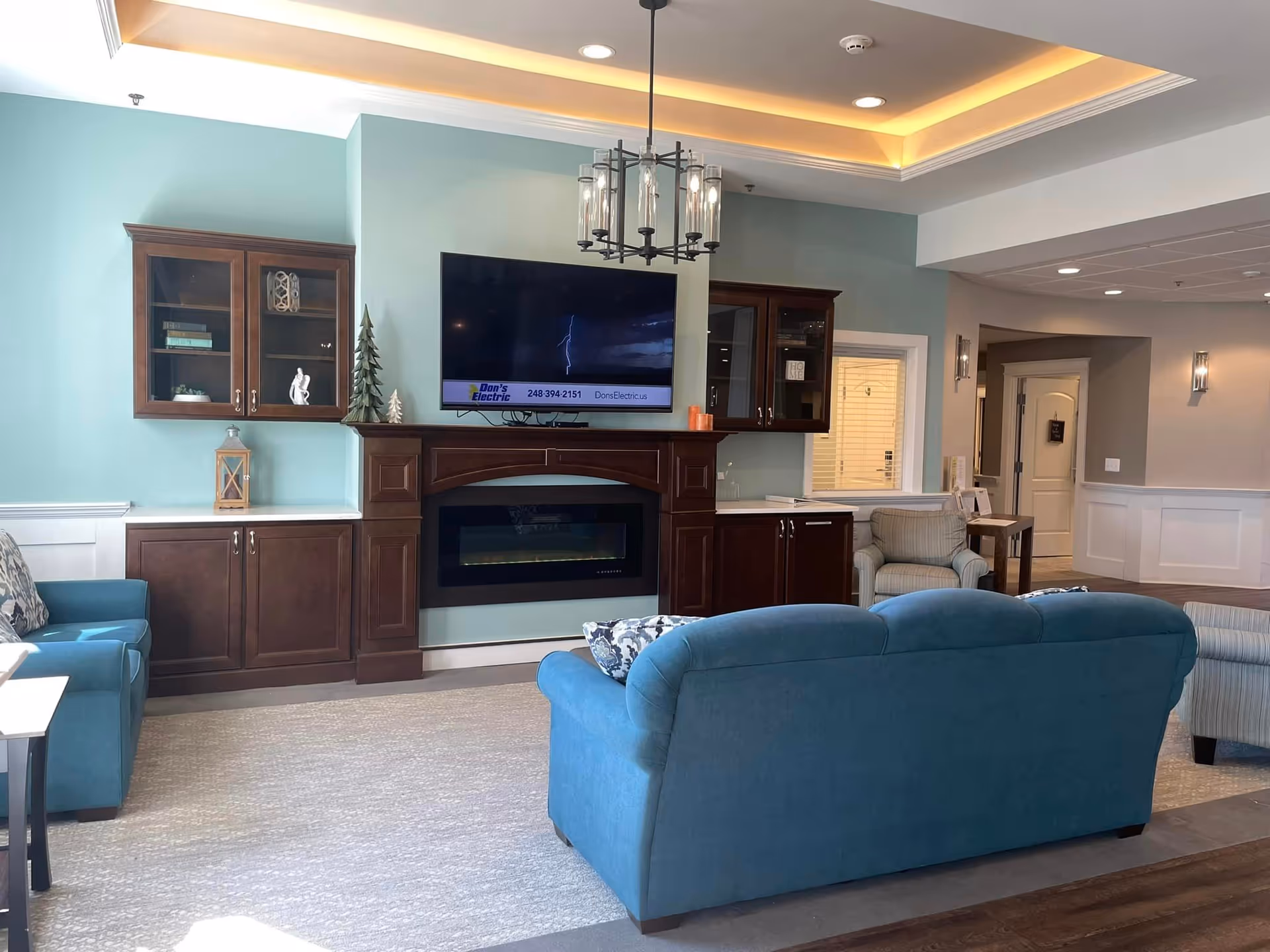A cozy senior living common area with teal walls and dark wood cabinetry. The room features a mounted flat-screen TV above a built-in electric fireplace, two blue upholstered sofas, a beige armchair, and a chandelier hanging from a tray ceiling with recessed lighting.