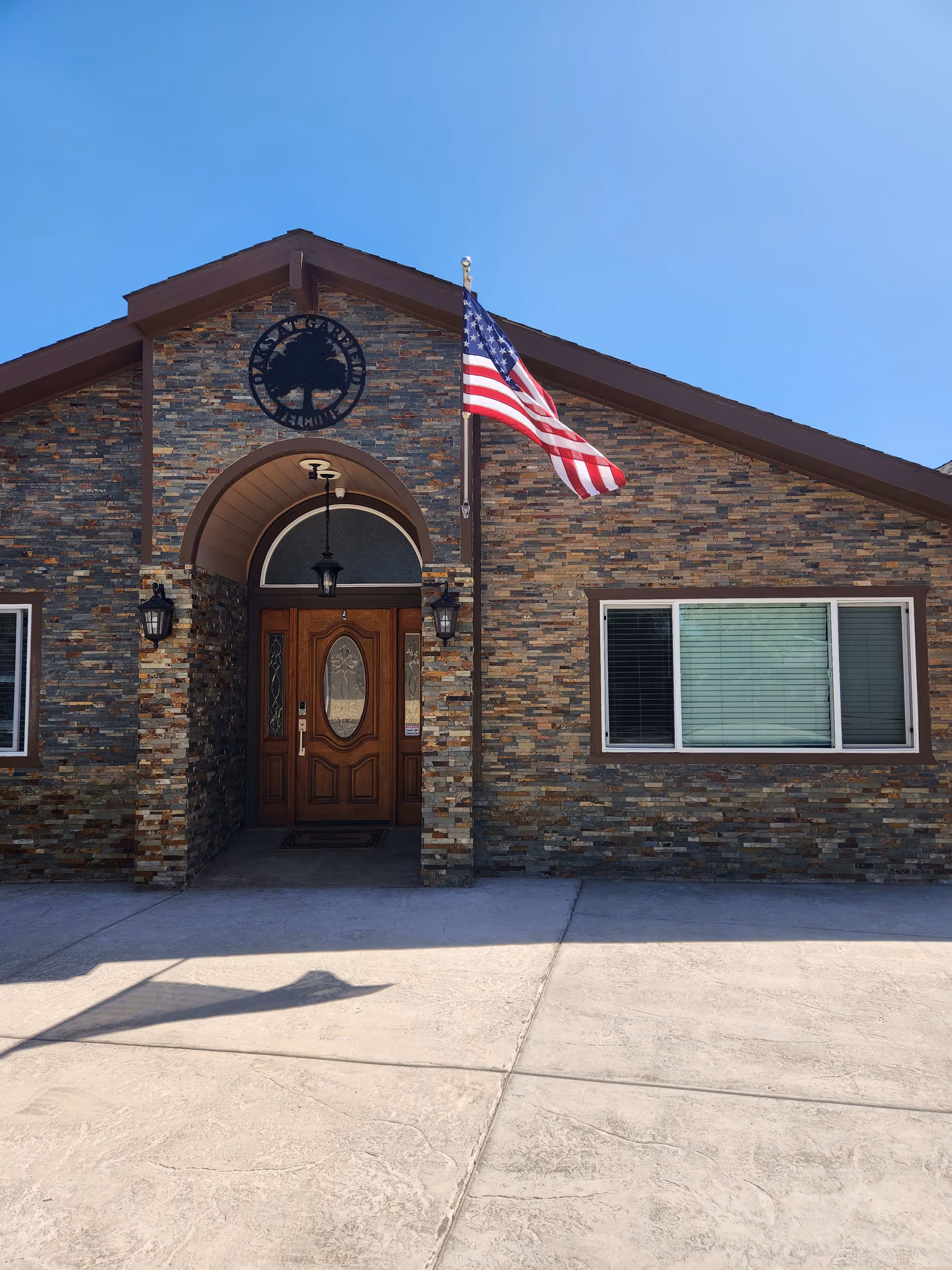 Front exterior view of a building with a stone facade, a wooden door with decorative glass, two wall lanterns on either side of the entrance, an American flag on a flagpole to the right of the door, and a clear blue sky above.