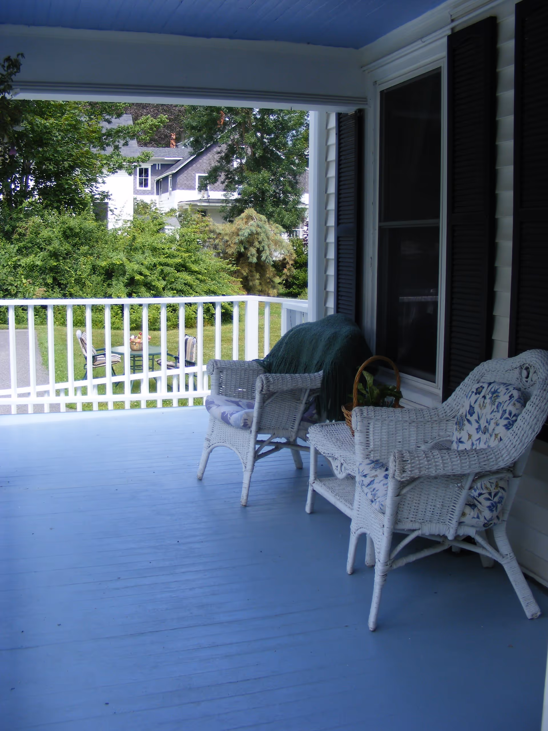 A covered blue porch with white wicker chairs and a railing overlooking a green yard and neighboring houses.