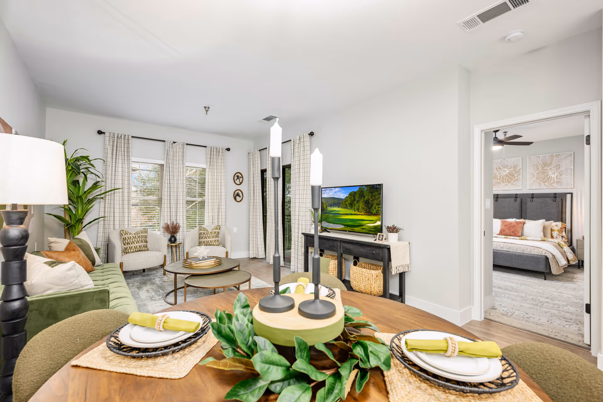 A bright and modern living room and dining area in a senior living facility. The dining table is set with plates, napkins, and a centerpiece with candles and greenery. The living room features a green sofa, two beige armchairs, a round coffee table, and a TV on a black console. Large windows with light-colored curtains allow natural light to fill the space. A doorway leads to a bedroom with a bed, pillows, and wall art.