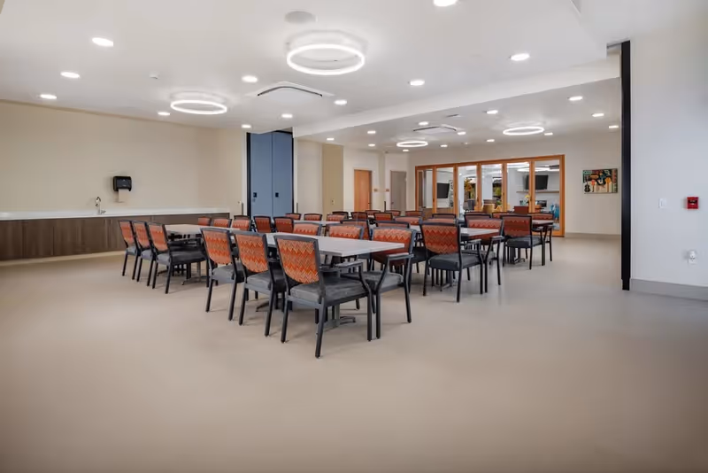 A spacious dining room with multiple rectangular tables and chairs arranged neatly. The chairs have patterned orange backs and dark seats. The room features modern circular ceiling lights, a beige floor, and a counter with a sink along one wall. There are large windows and doors in the background, allowing natural light to enter.