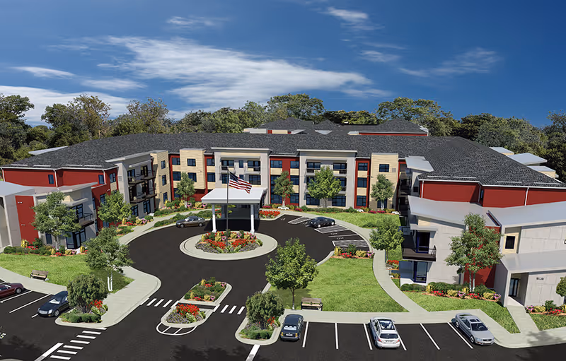 Aerial view of a senior living facility building with a circular driveway and landscaped gardens. The building has multiple stories with red and beige exterior walls, surrounded by trees and parking spaces with several cars parked.