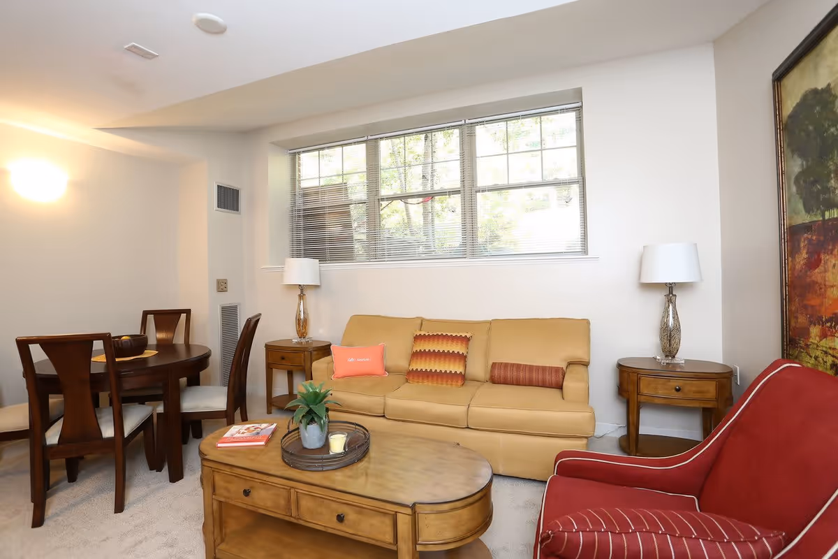 A cozy living room area with a beige sofa adorned with three decorative pillows, a red armchair with white piping, and a wooden coffee table with a plant, candle, and book on it. Behind the sofa is a large window with blinds partially open, letting in natural light. To the left, there is a wooden dining table with four chairs, and two matching wooden side tables with lamps on either side of the sofa. A large framed painting is visible on the right wall.