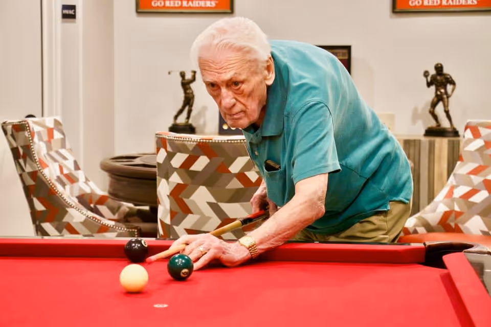 An elderly man in a teal polo shirt playing pool on a red pool table in a room with patterned chairs and sports trophies in the background.