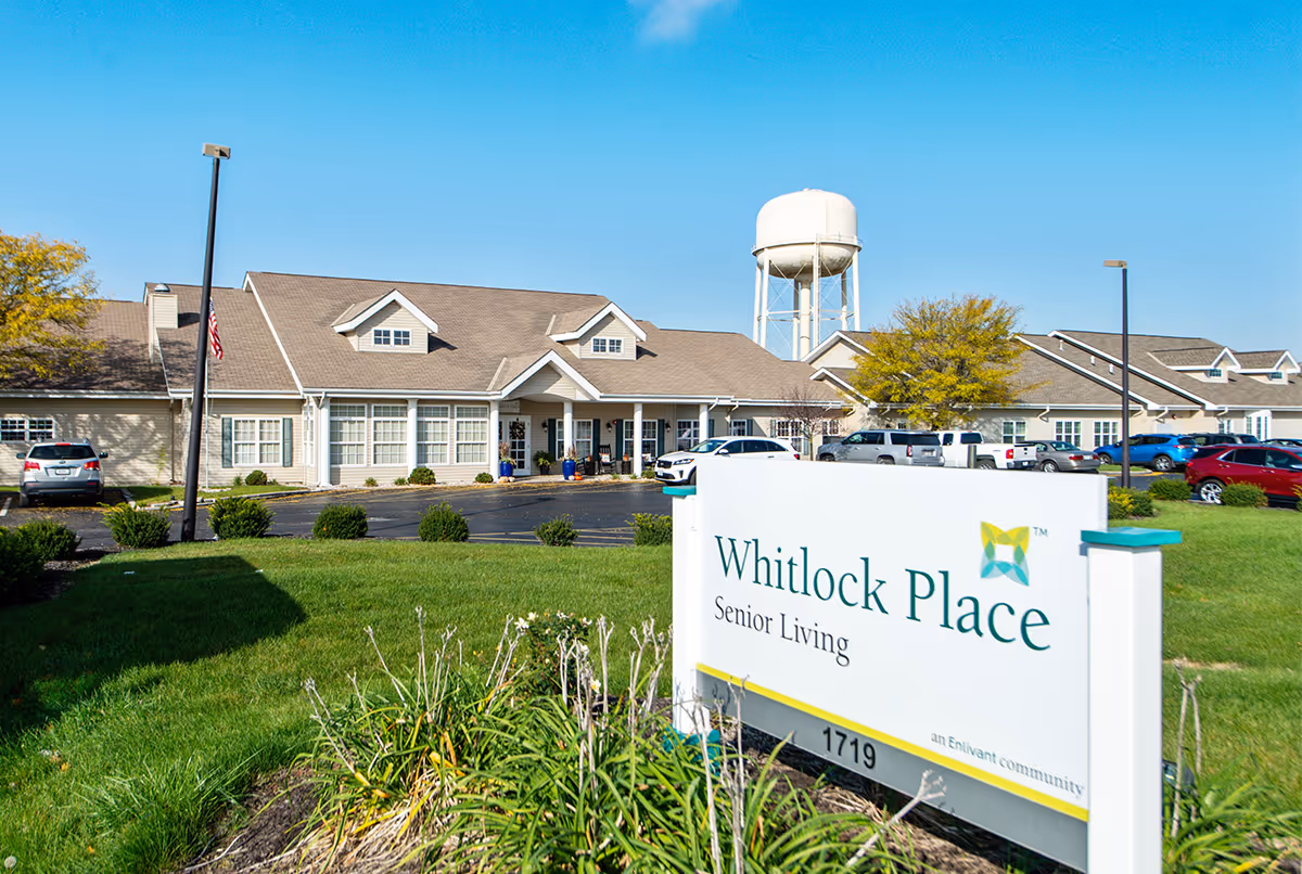 Exterior view of Whitlock Place Senior Living facility with a clear blue sky, a water tower in the background, a parking lot with several cars, and a sign in the foreground displaying the facility name and address.