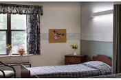 A simple, clean nursing home bedroom with a single bed covered in a checkered blanket. There is a wooden nightstand with a small vase of flowers next to the bed. A window with floral curtains lets in natural light, and a butterfly-themed picture hangs on the wall above the nightstand.