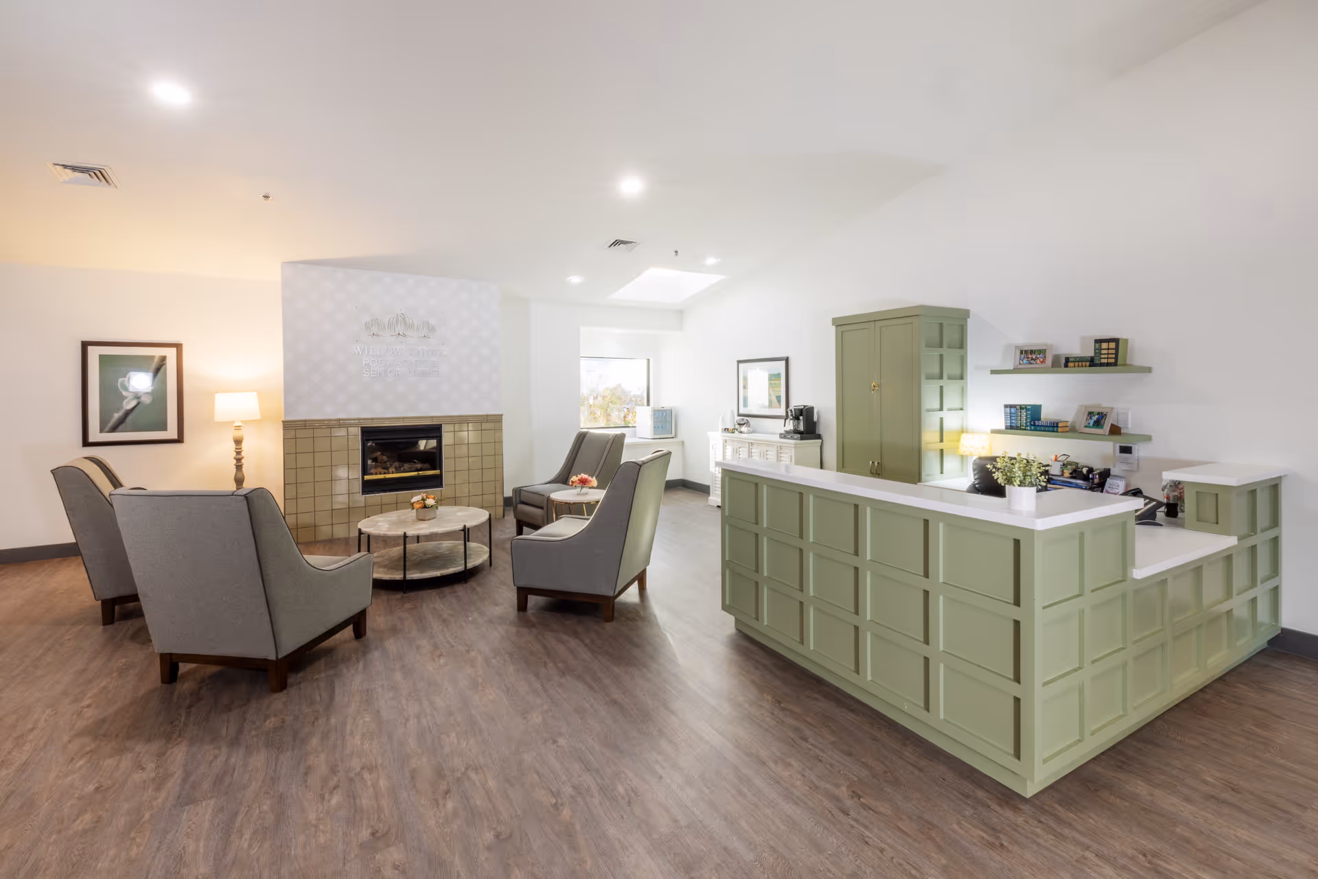 A bright and spacious senior living facility common area featuring a green reception desk on the right, four gray armchairs arranged around two round coffee tables in front of a fireplace with a tiled surround, a floor lamp, framed artwork on the wall, and shelves with decorative items and office supplies.