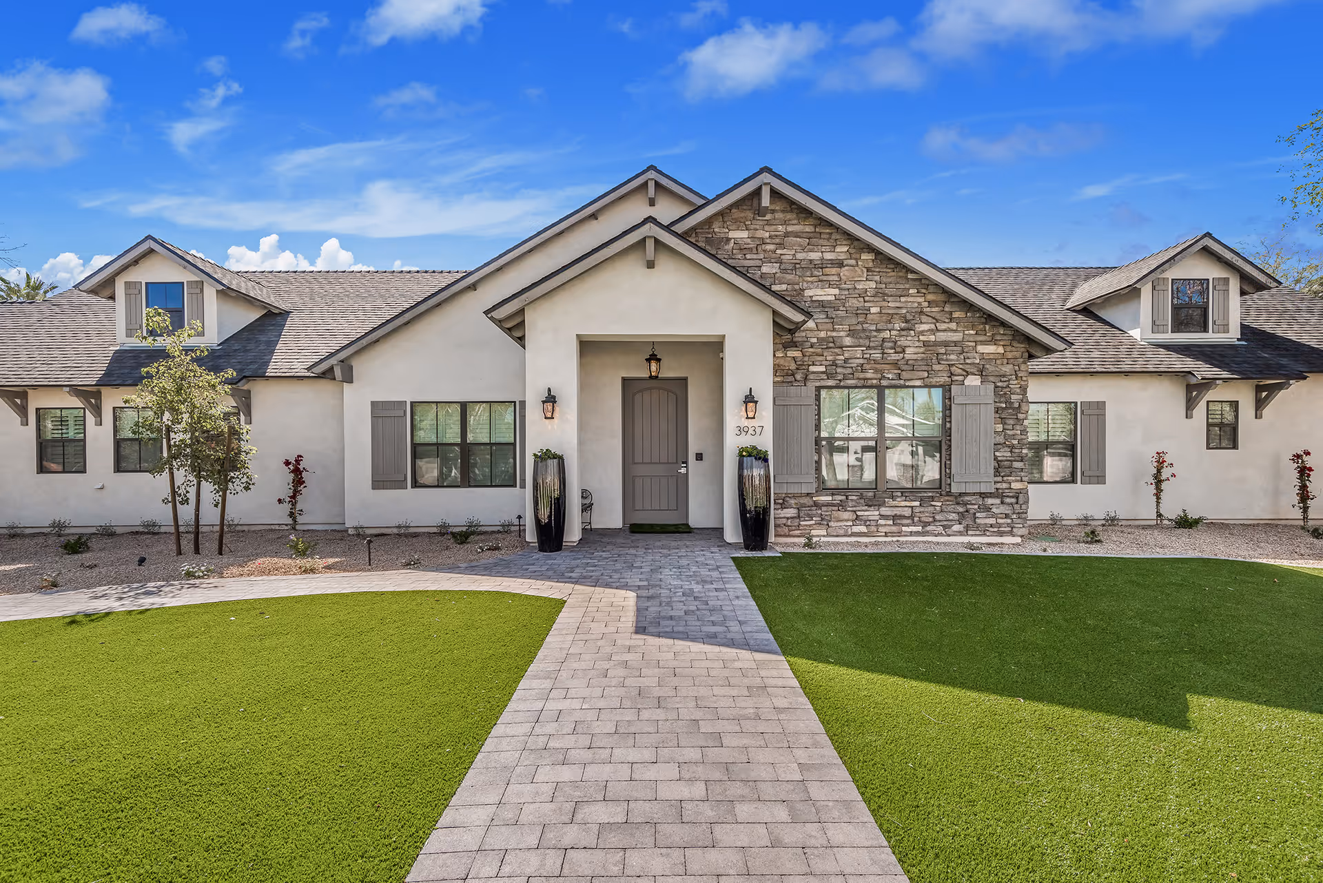 Front exterior view of a single-story building with a combination of stone and stucco facade, a central entrance with a gray door, two tall planters on either side, multiple windows with shutters, and a paved walkway leading to the entrance surrounded by green lawn.
