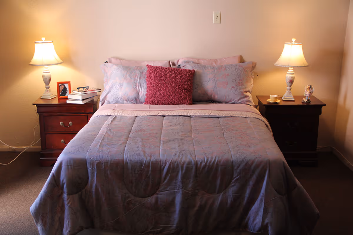 A neatly made bed with a purple and pink floral comforter and pillows, including a textured dark pink accent pillow in the center. On each side of the bed are wooden nightstands with matching table lamps. The left nightstand has a framed photo, a pair of glasses, and a stack of books. The right nightstand has a small cup and saucer and a decorative figurine. The room has beige walls and carpeted floor.