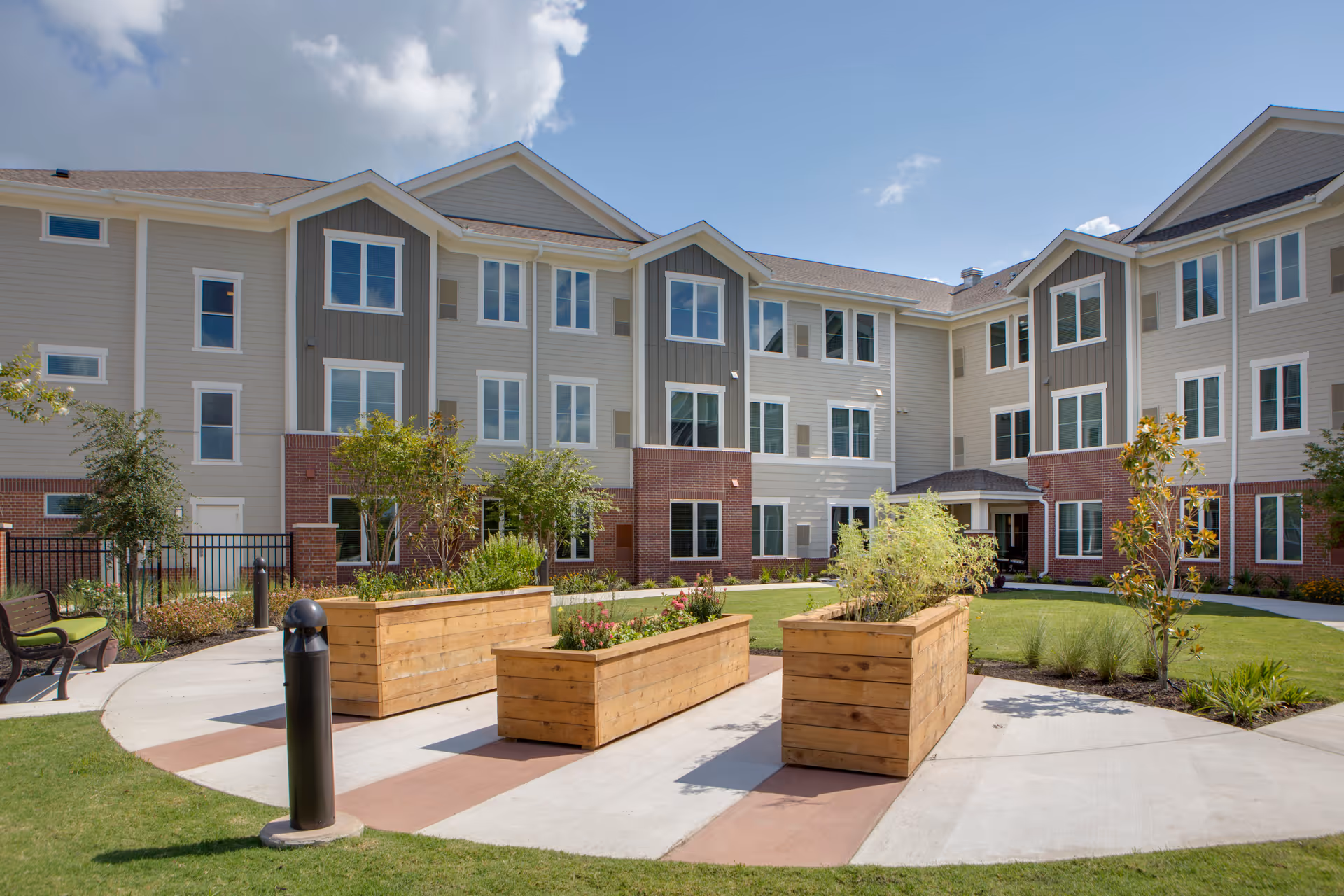 Outdoor courtyard area of Delaney Parkway Lakes facility with a three-story building in the background. The courtyard features a paved circular walkway, wooden planter boxes with plants, green grass, small trees, and benches under a partly cloudy sky.