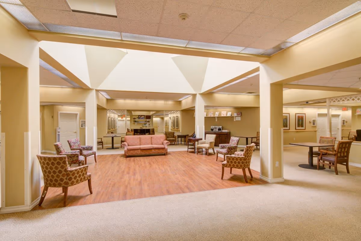 A spacious senior living facility common area with a central seating arrangement including a pink sofa and several patterned armchairs on a wooden floor. The room has beige walls, a carpeted floor surrounding the wooden area, and a ceiling with recessed lighting. There are tables and chairs along the perimeter and framed artwork on the walls.