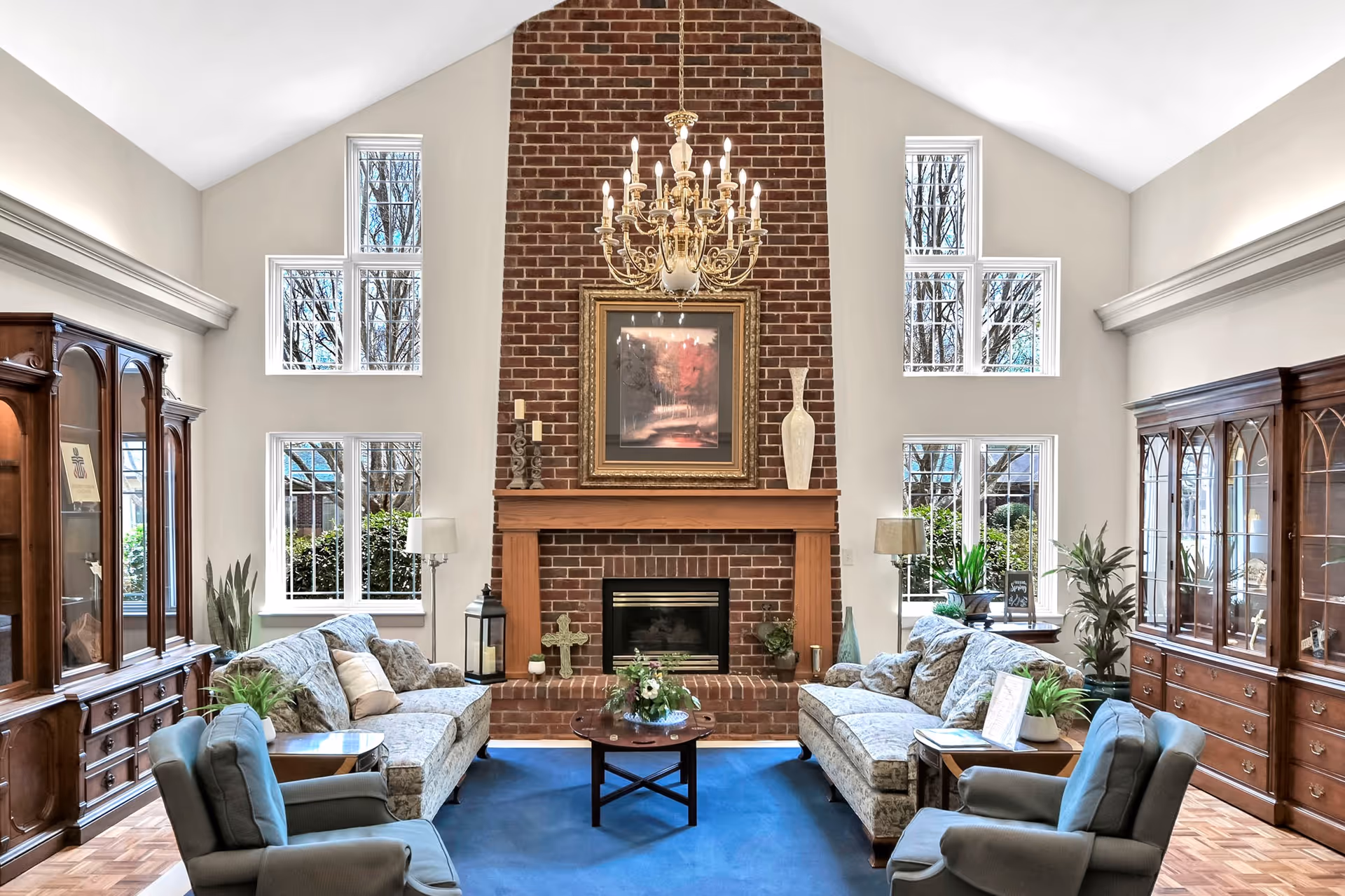 Spacious communal living room with high vaulted ceiling, central brick fireplace and chandelier, sofas and armchairs arranged around a coffee table.