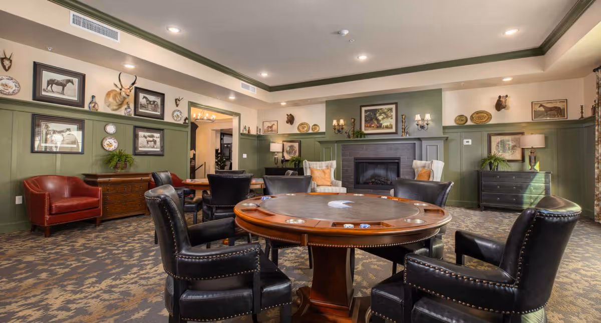 Cozy common room with a round gaming table surrounded by leather chairs, a fireplace and framed artwork on green paneled walls.