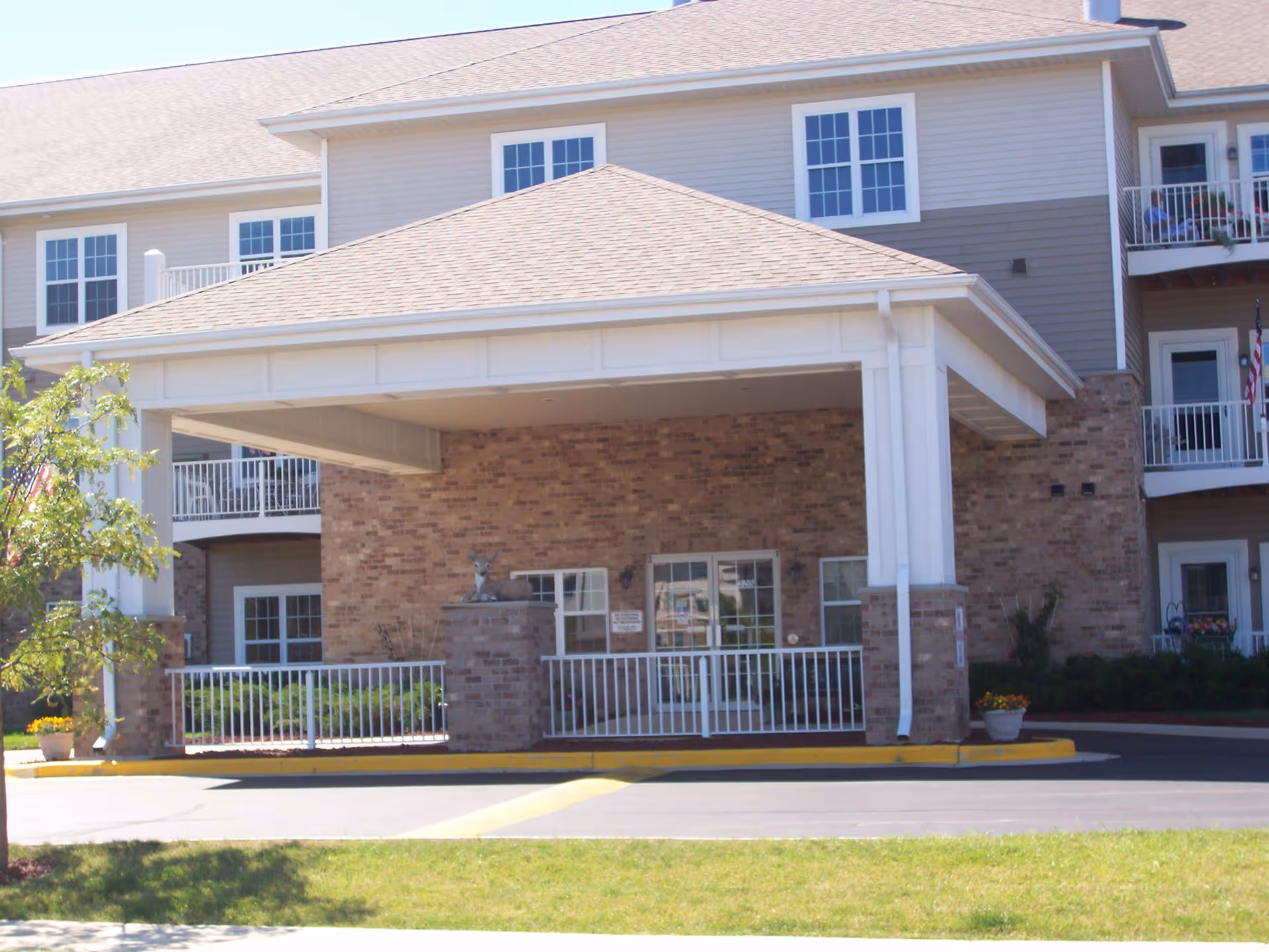 Entrance of a senior living facility with a covered drop-off area supported by white columns and brick pillars. The building has beige siding and multiple windows, with balconies visible on the right side. There is a small statue of a deer on one of the brick pillars near the entrance.