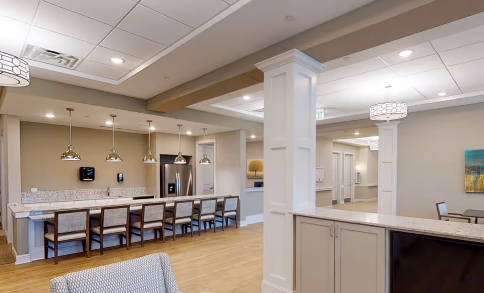Interior view of a senior living facility kitchen and common area with a long counter and seven chairs, pendant lights hanging above the counter, stainless steel refrigerator, and light-colored walls and flooring.