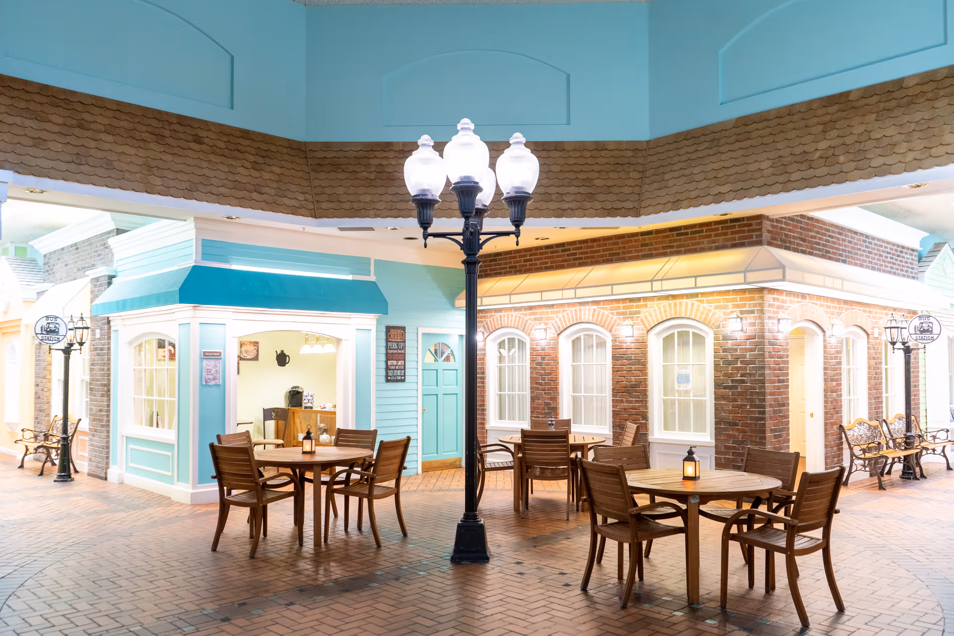 Indoor seating area with wooden tables and chairs arranged around a black street lamp. The surrounding walls are designed to look like small storefronts with windows and doors, featuring pastel blue and brick facades. Benches and decorative street lamps line the walkway, creating a cozy, village-like atmosphere inside a senior living facility.