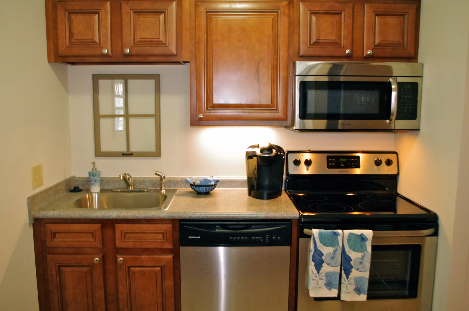 A small kitchen area with wooden cabinets, a stainless steel microwave, electric stove, dishwasher, and a sink. There is a coffee maker on the counter and two blue patterned towels hanging on the oven handle.